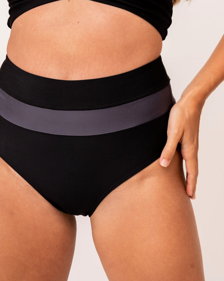 A studio image of a women wearing black and grey color blocked high waisted swimsuit bottoms.